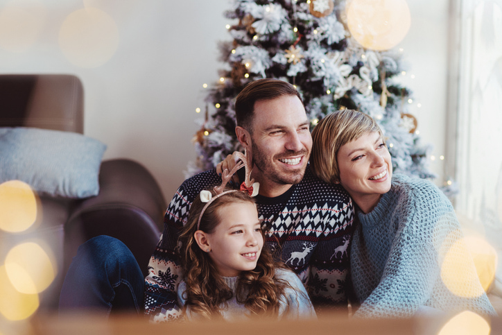 Father and mother with young girl in front of a Christmas tree, smiling and holding each other